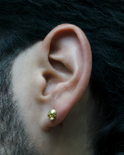 A solid yellow gold skull stud earring. Shown on an ear from the front.