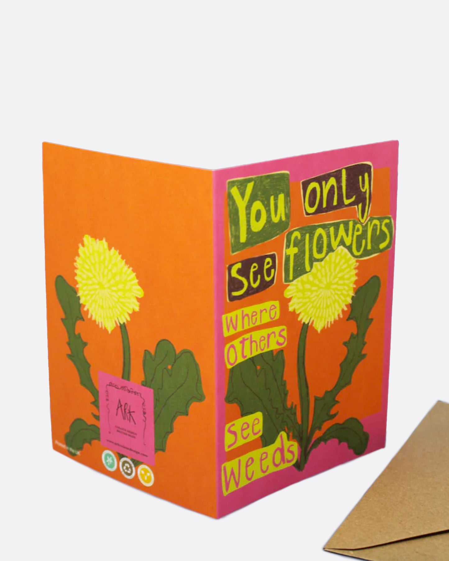 A bright pink and orange greeting card with a dandelion on the front and the words 'you only see flowers where other see weeds,' and a blank white interior. Shown with its kraft paper envelope.