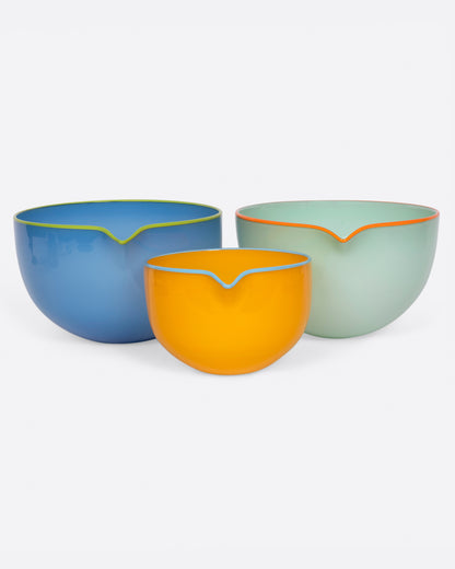 A set of three hand blown glass spouted pouring bowls- two large and one small. The large bowls are blue with a chartreuse edge and green with an orange edge. The small bowl is yellow with a blue edge.  Shown from the front.