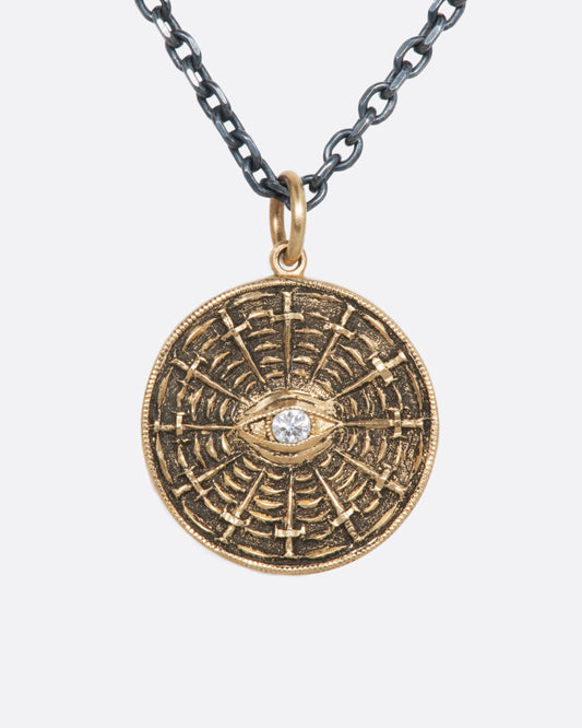 An oxidized sterling silver necklace with a yellow disc pendant, with a diamond eye at its center and swords radiating around it. 