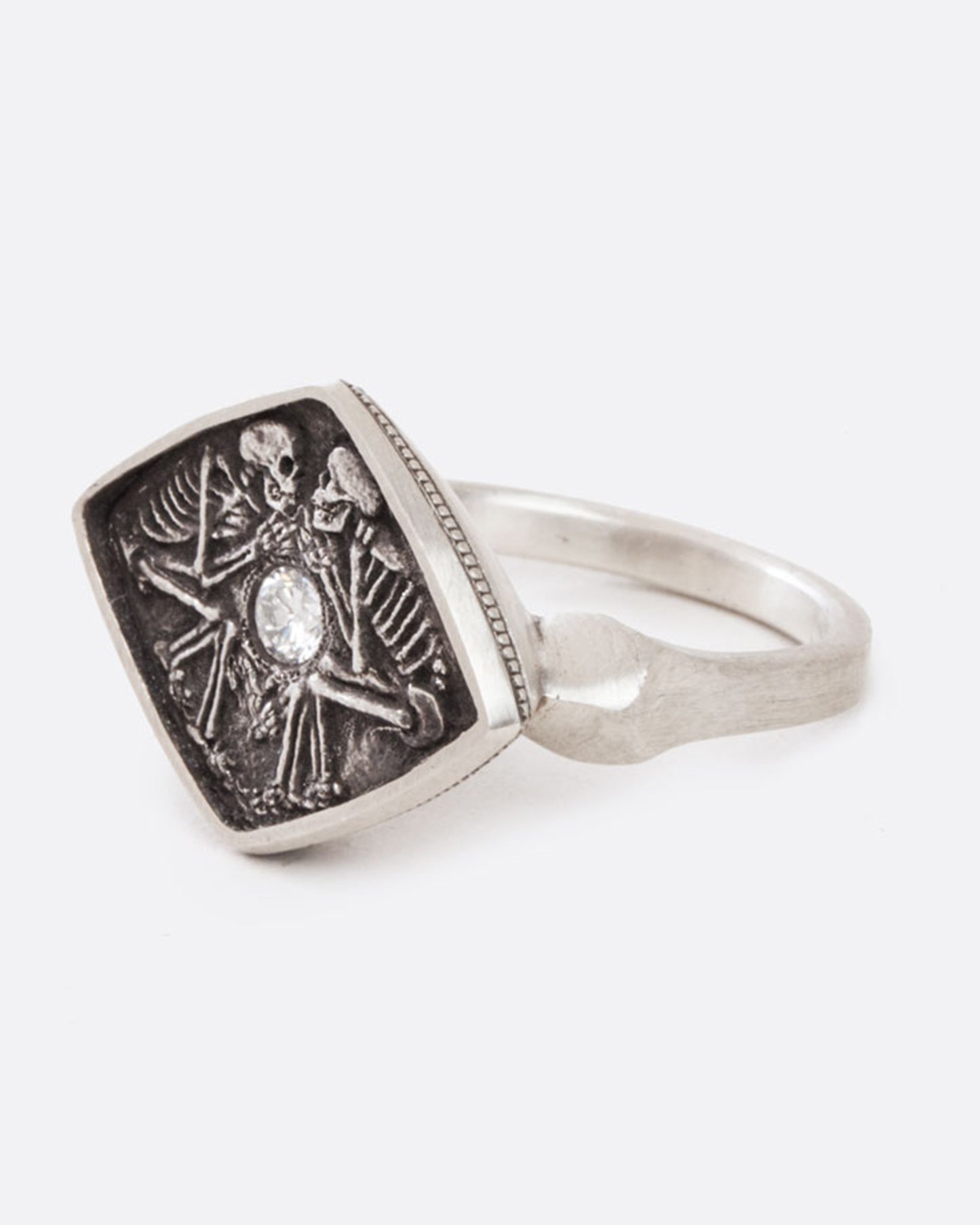 A sterling silver ring with two skeletons facing each other, lying in lovely death with a round diamond resting in their hands, glowing between them