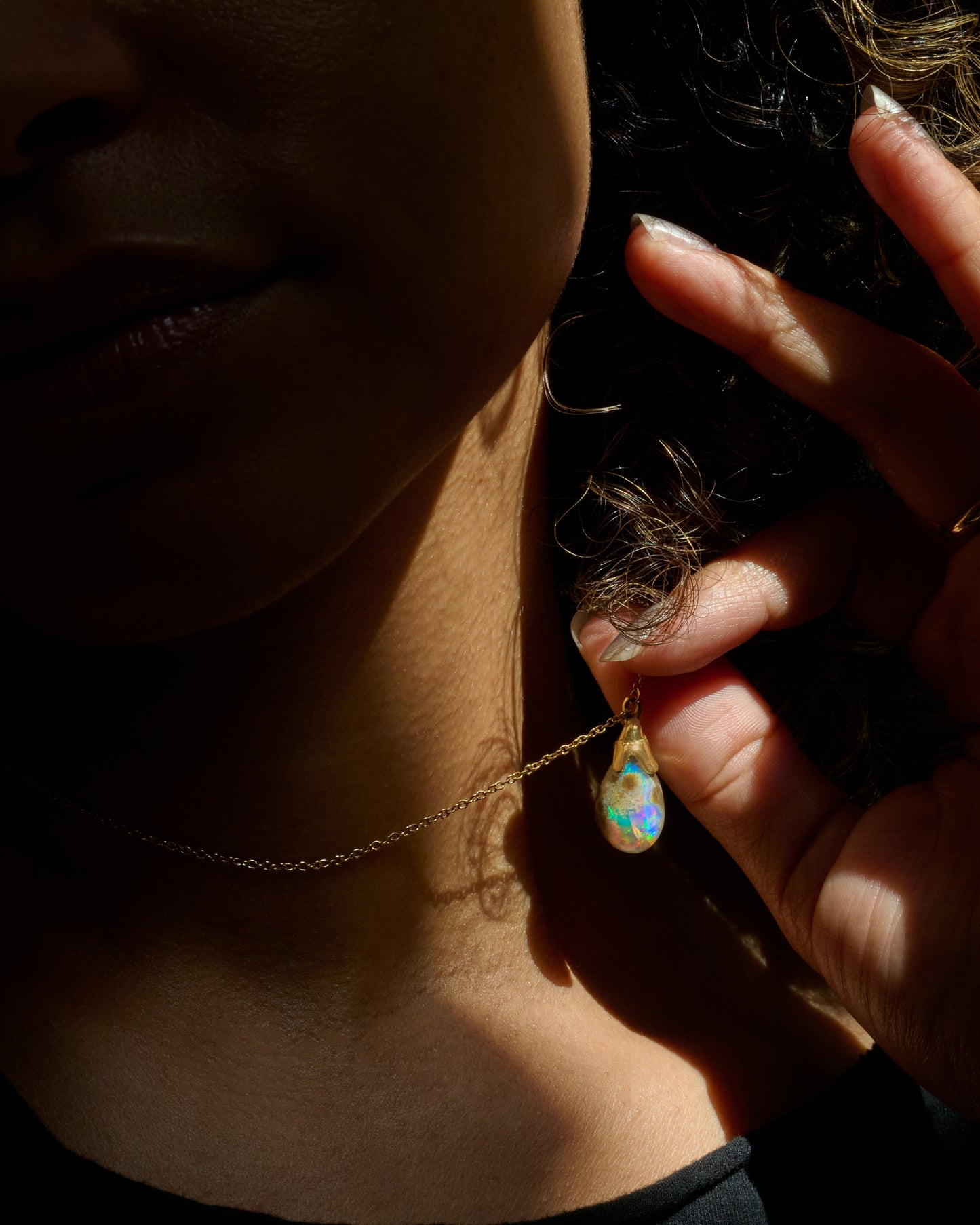 A close-up lifestyle shot of a person wearing a delicate gold chain necklace with a teardrop-shaped iridescent opal pendant, held by a hand in dramatic, high-contrast sunlight.