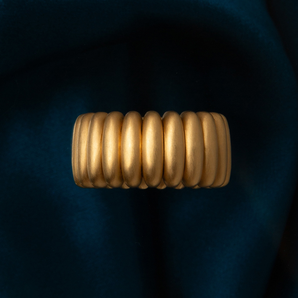 A wide, ribbed gold ring with a textured, segmented design, displayed against a dark navy blue velvet fabric background.