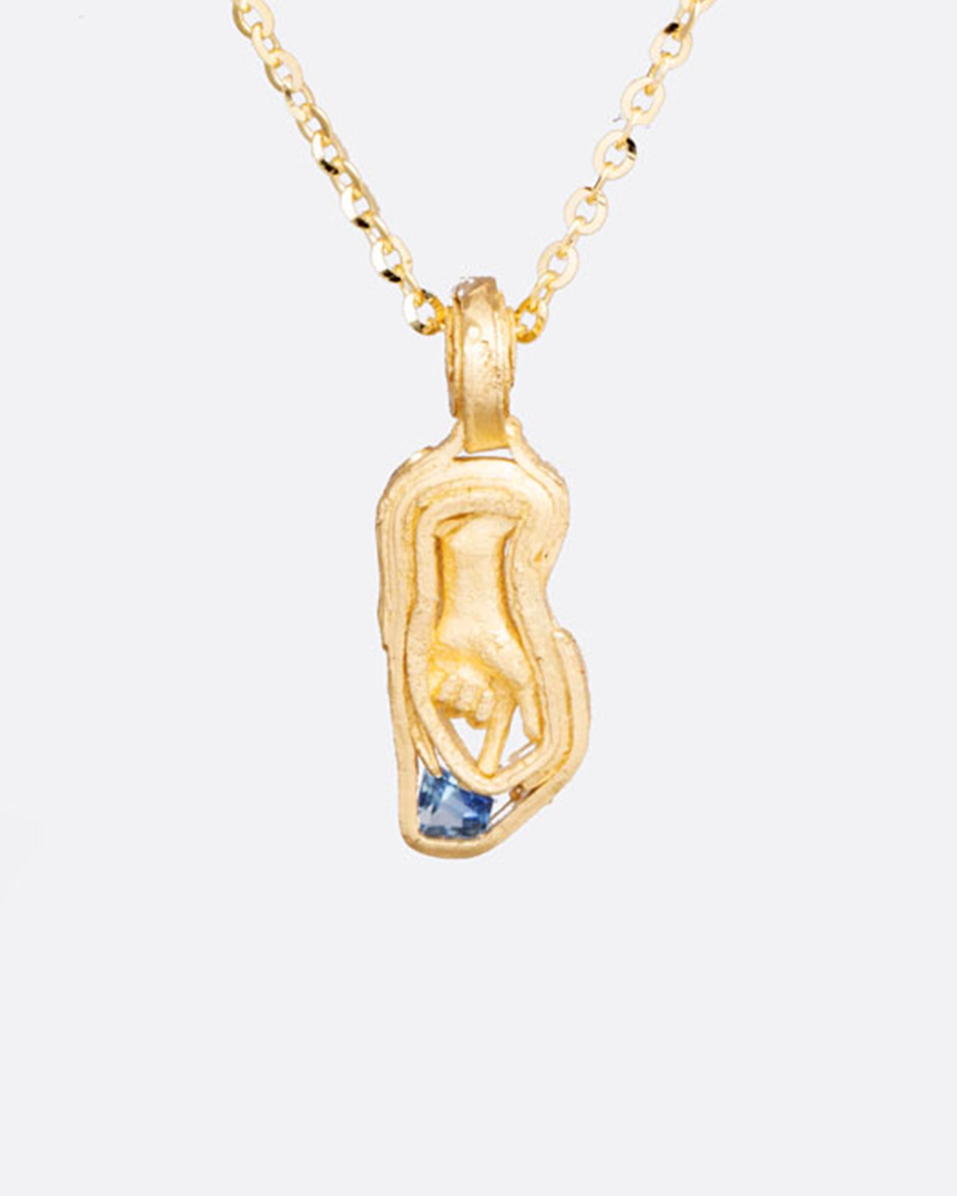 An intricate sculptural hand is surrounded by wrapped yellow gold. Its finger points, directing the eye, to the small blue sapphire at the tip of the pendant.