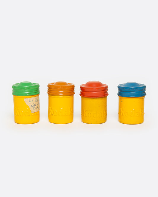 Four Kodak Color Linen film canisters in a row, shown from the front. Each yellow canister has a different colored lid: green, orange, red, and blue.