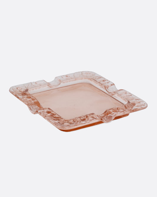 A square pink glass ashtray with a floral design on the edges, shown from the side.