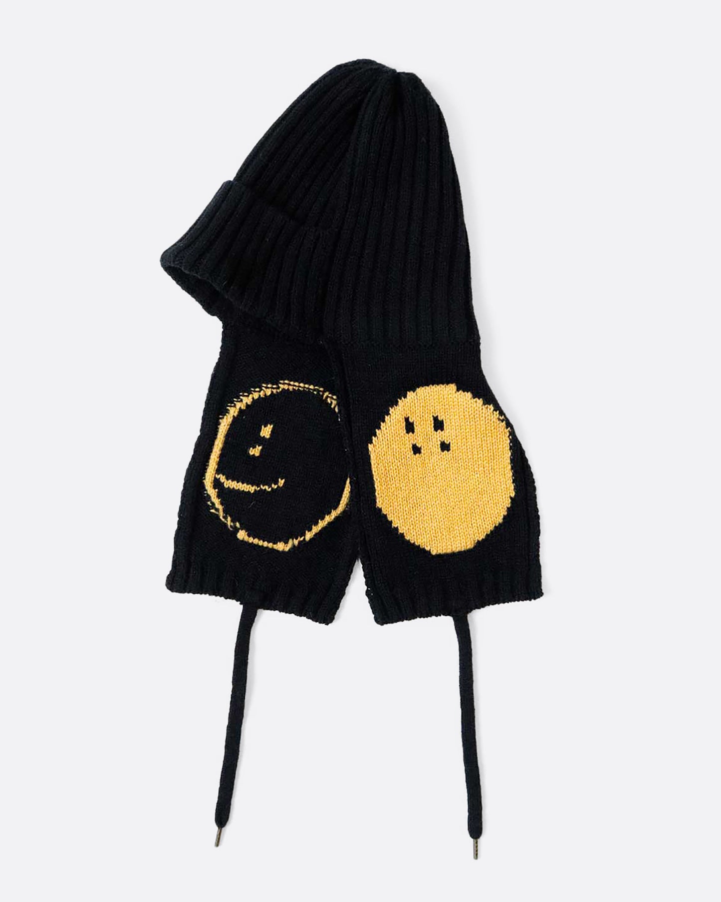 A black knit hat with yellow smilie faces on either ear flap. View from the side, laying flat.