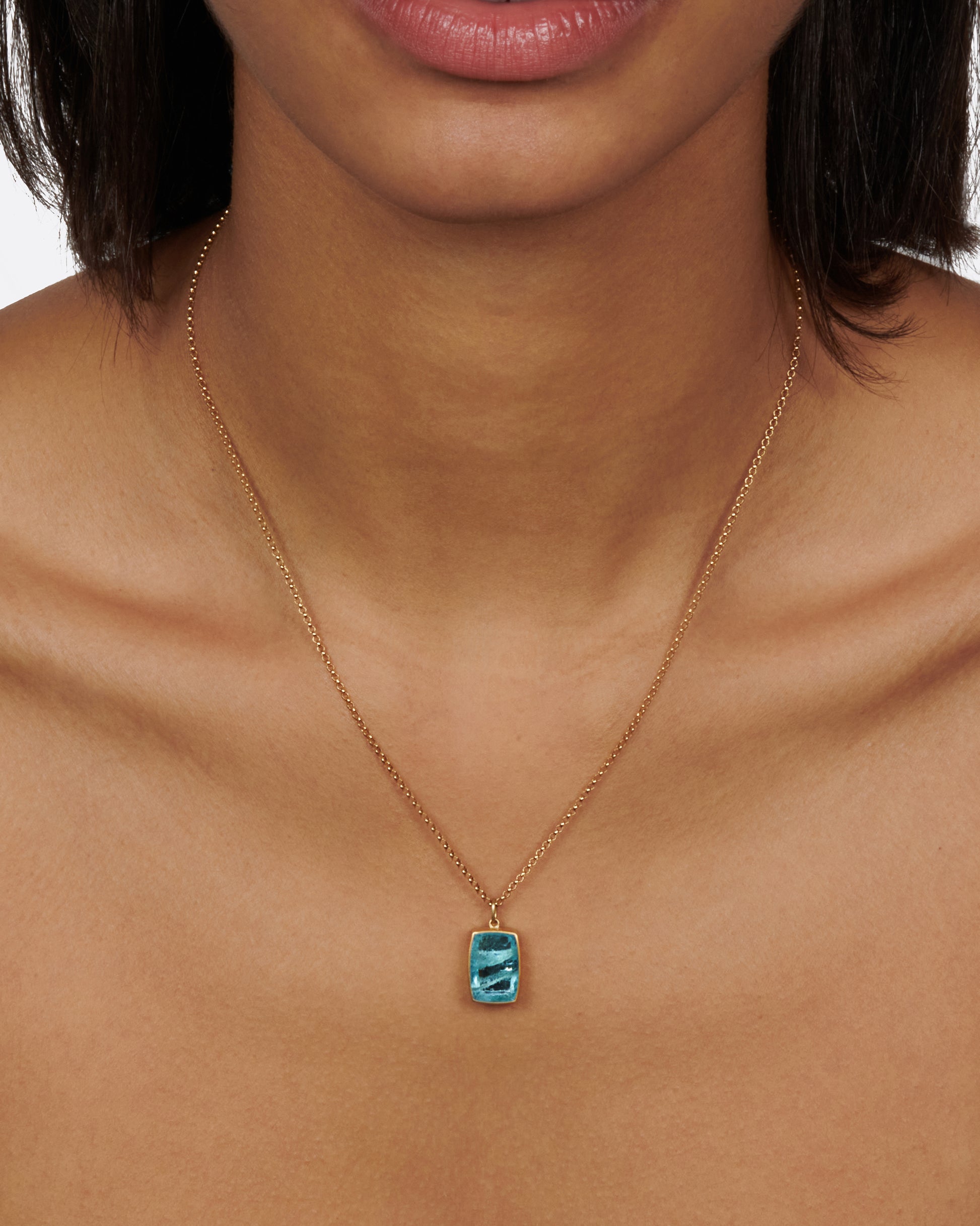 A zoomed out view of a cushion shaped aquamarine pendant with a yellow gold bezel on a gold chain on a neck..