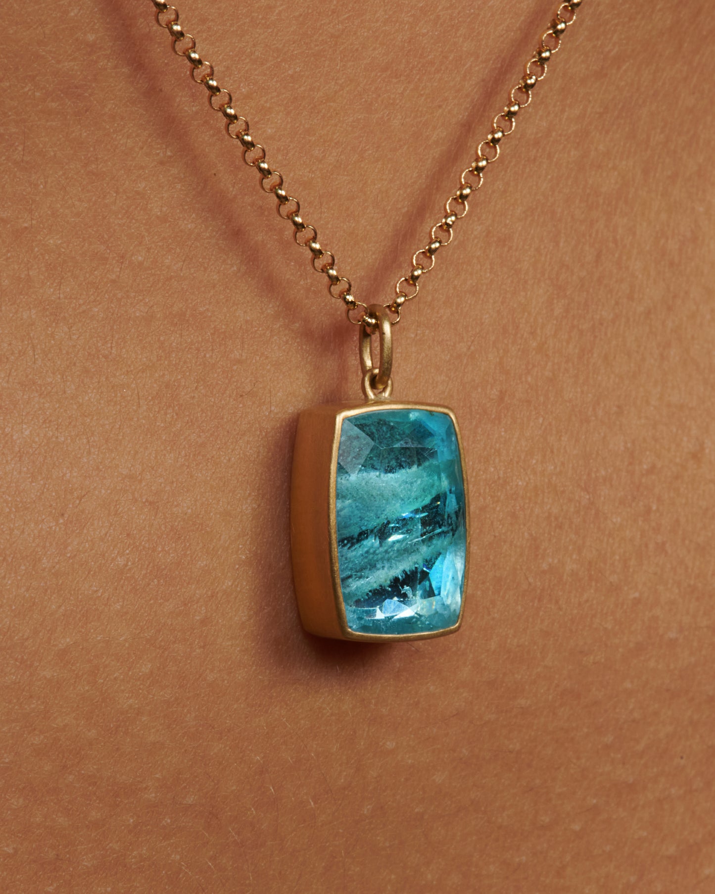 A close up of a cushion shaped aquamarine pendant with a yellow gold bezel on a gold chain on a neck.