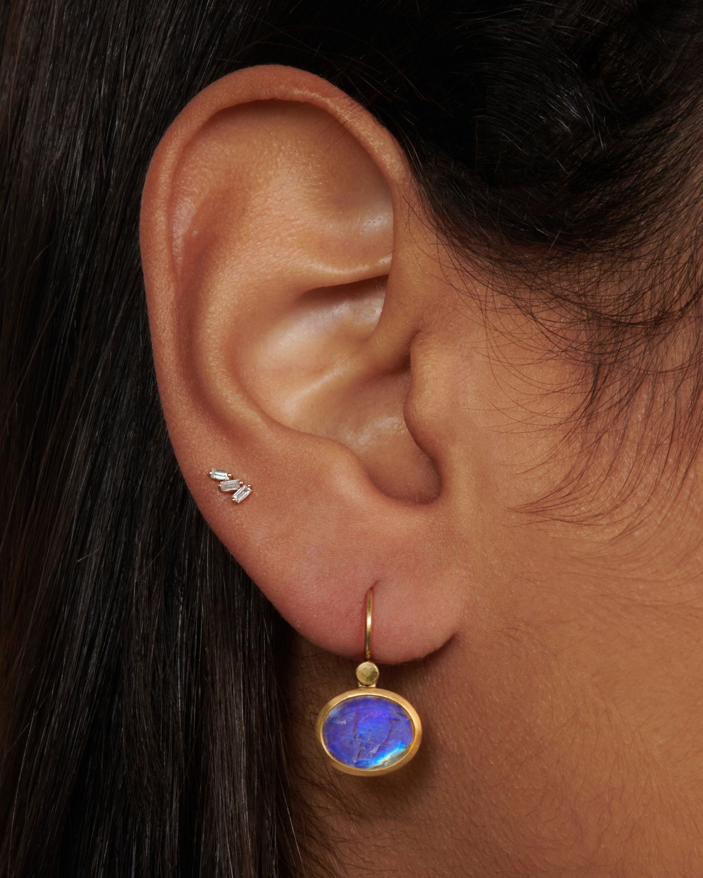 An oval moonstone earring set in a yellow gold bezel on an ear.