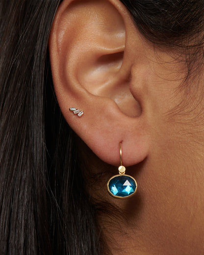 Lola Brooks earring in 18k yellow gold worn on an ear, a small faceted oval London blue topaz in a gold bezel on a hook wire