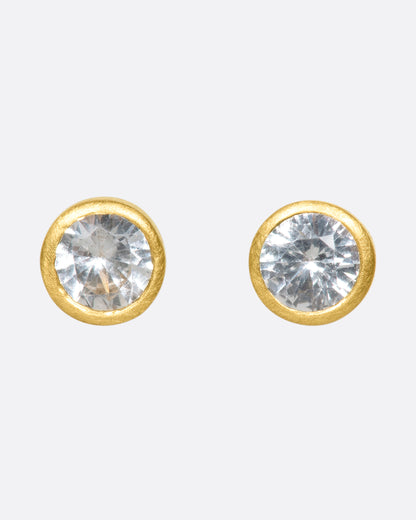 A pair of yellow gold solitaire earrings with round white sapphires in bezel settings.