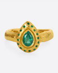 A high karat gold ring with an organic shaped band and a pear shaped emerald at its center with a sunken round emerald halo.