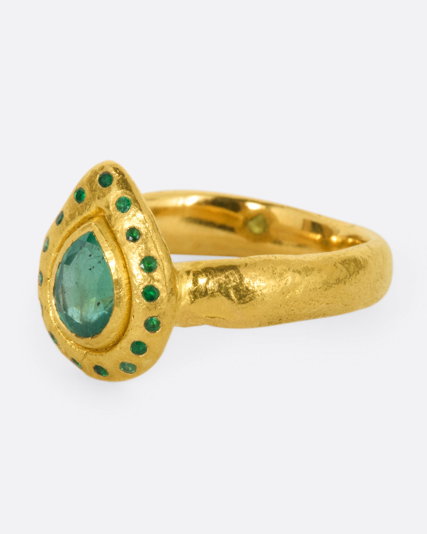 A high karat gold ring with an organic shaped band and a pear shaped emerald at its center with a sunken round emerald halo. Shown from the side.