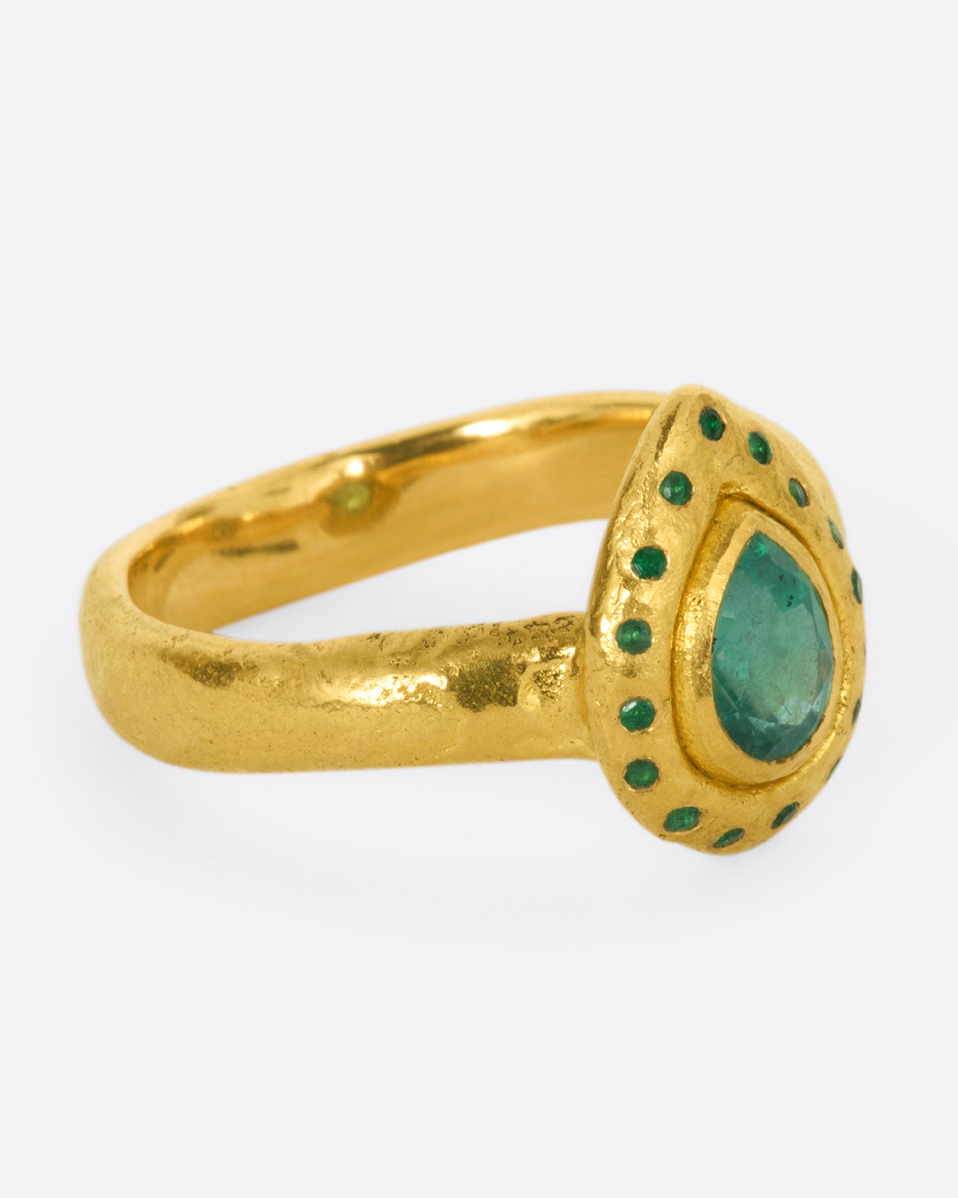 A high karat gold ring with an organic shaped band and a pear shaped emerald at its center with a sunken round emerald halo. Shown from the side.