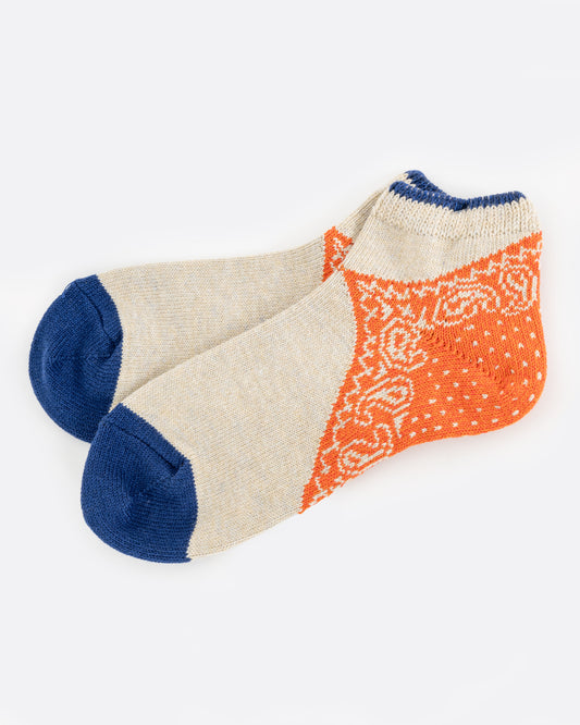 A pair of tan and blue ankle socks with an orange paisley bandana print heel. Shown lying flat from the side.