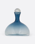 A blown glass decanter in a steel blue gradient with a wide base and a round silver stopper, shown from the front.