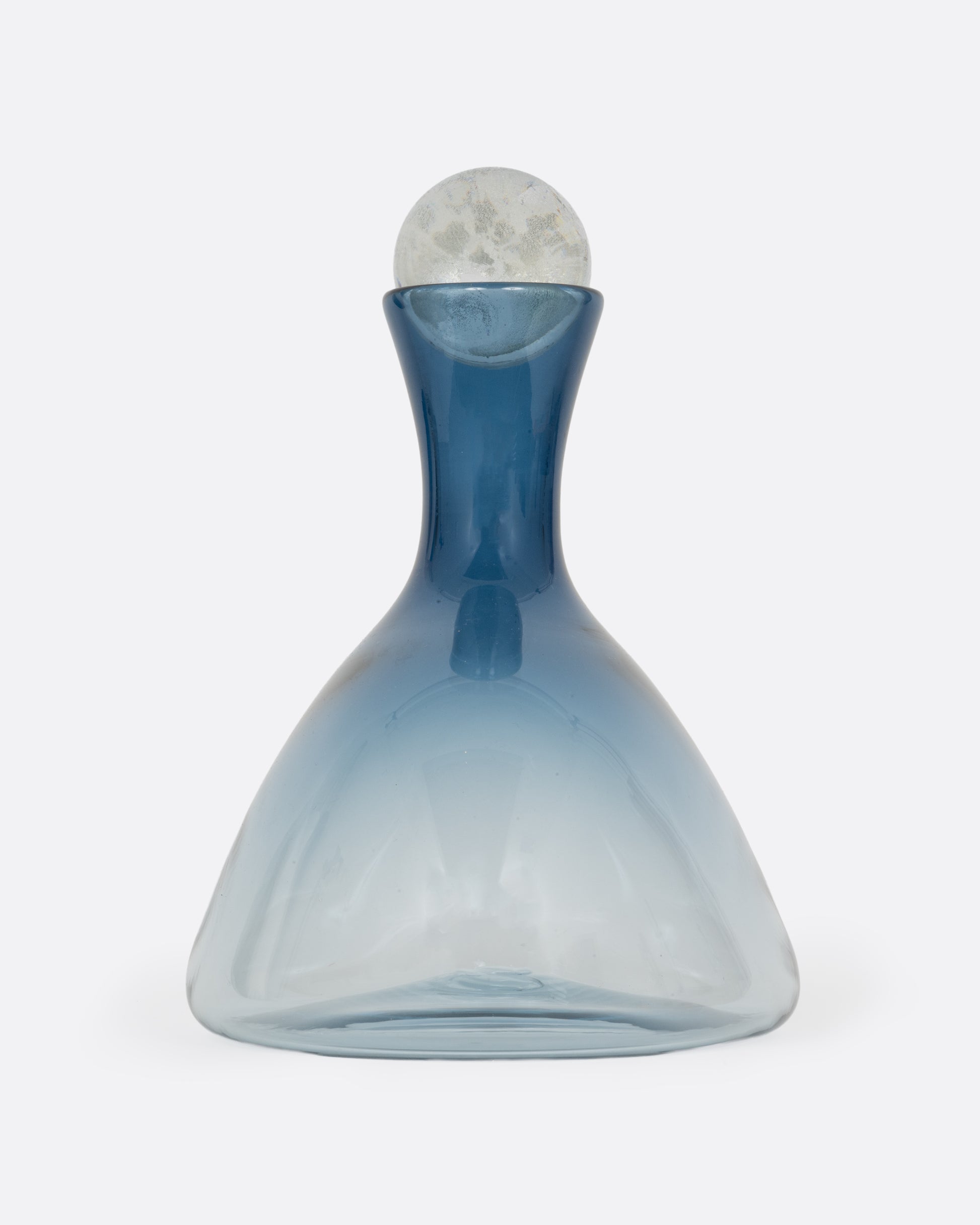 A blown glass decanter in a steel blue gradient with a wide base and a round silver stopper, shown from the side.