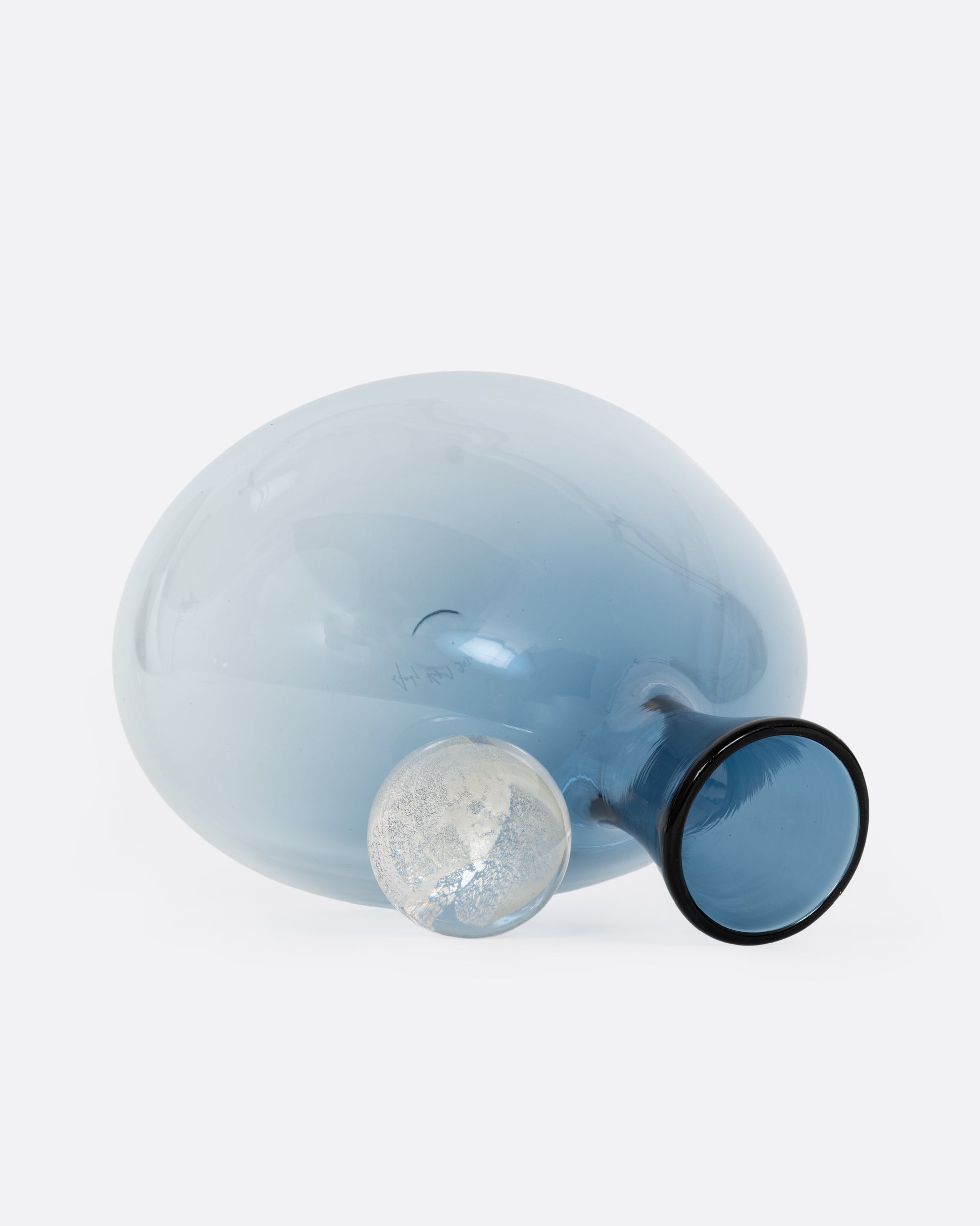 A blown glass decanter in a steel blue gradient with a wide base and a round silver stopper, shown on its side open.