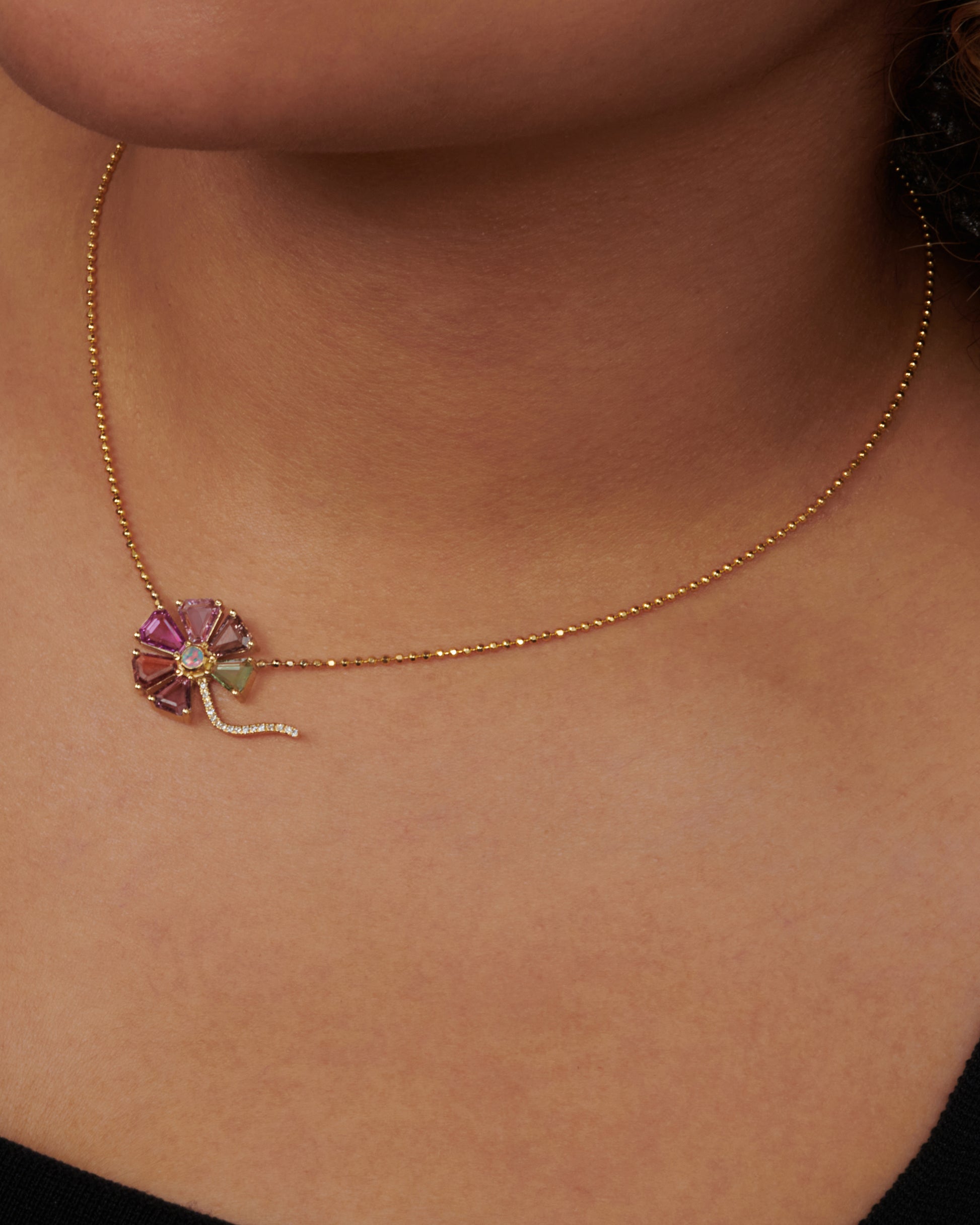 A zoomed out view of a flower pendant with sapphire petals, diamond stem, and opal center on a gold ball chain. Shown on a neck.