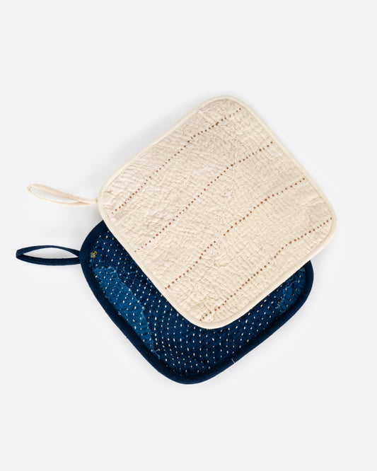 A pair of pot holders- one cream and one indigo blue. Both have contrast stitching. Shown from the front stacked.