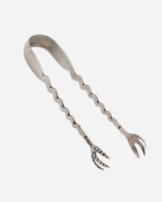 A pair of silver tongs with an arched top, zigzag arms, and claw like 'hands', shown from the side.