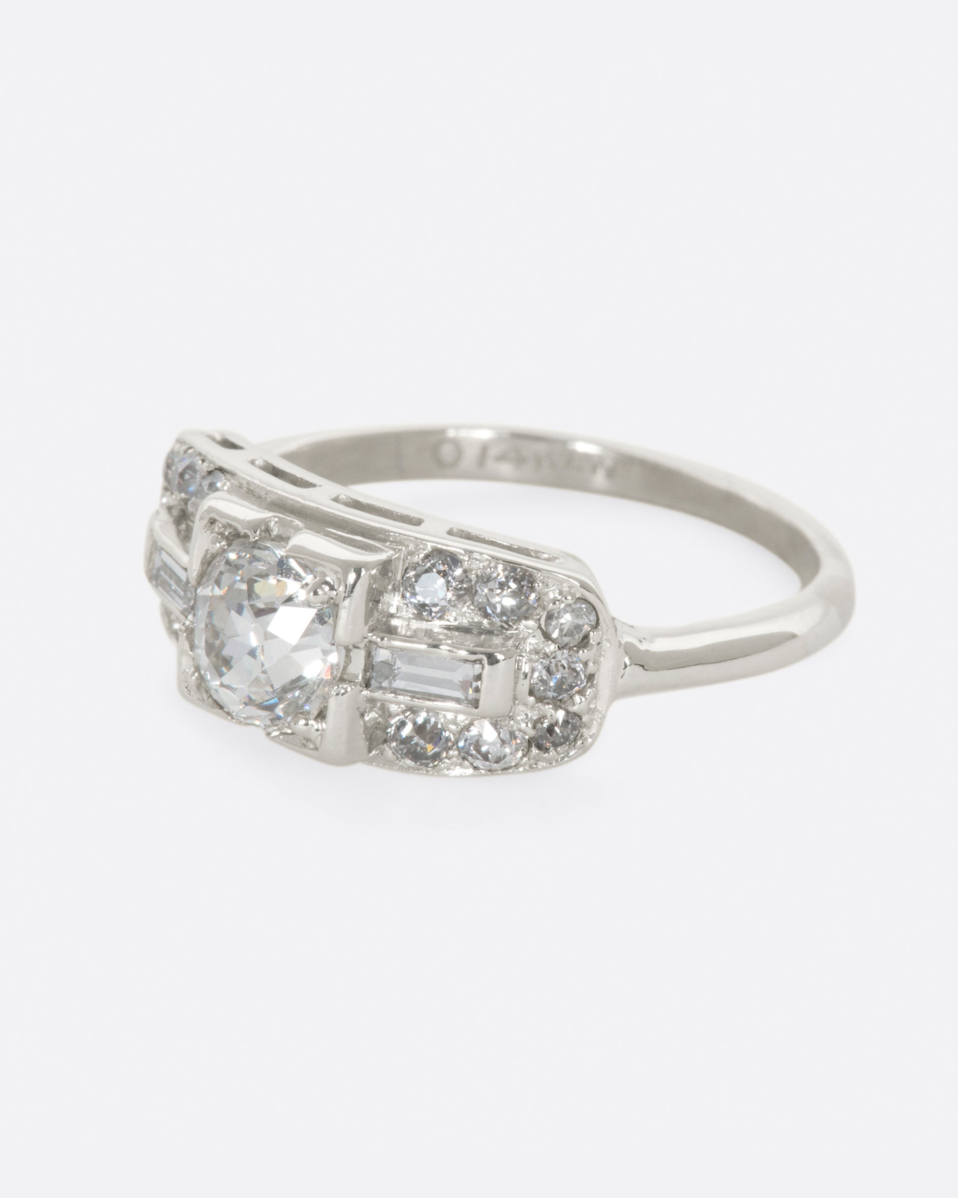 A white gold ring with round center diamond and baguette and round diamonds surrounding on a wide flat face. View from the left side.