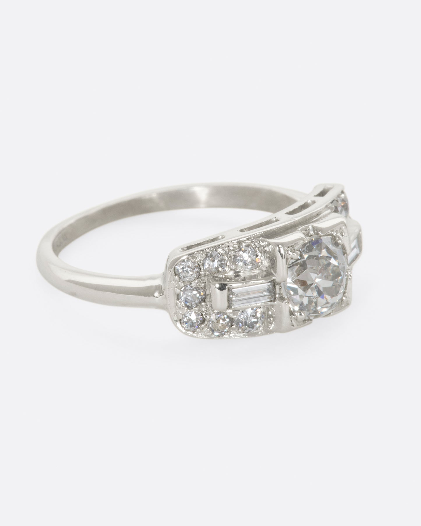 A white gold ring with round center diamond and baguette and round diamonds surrounding on a wide flat face. View from the right side.