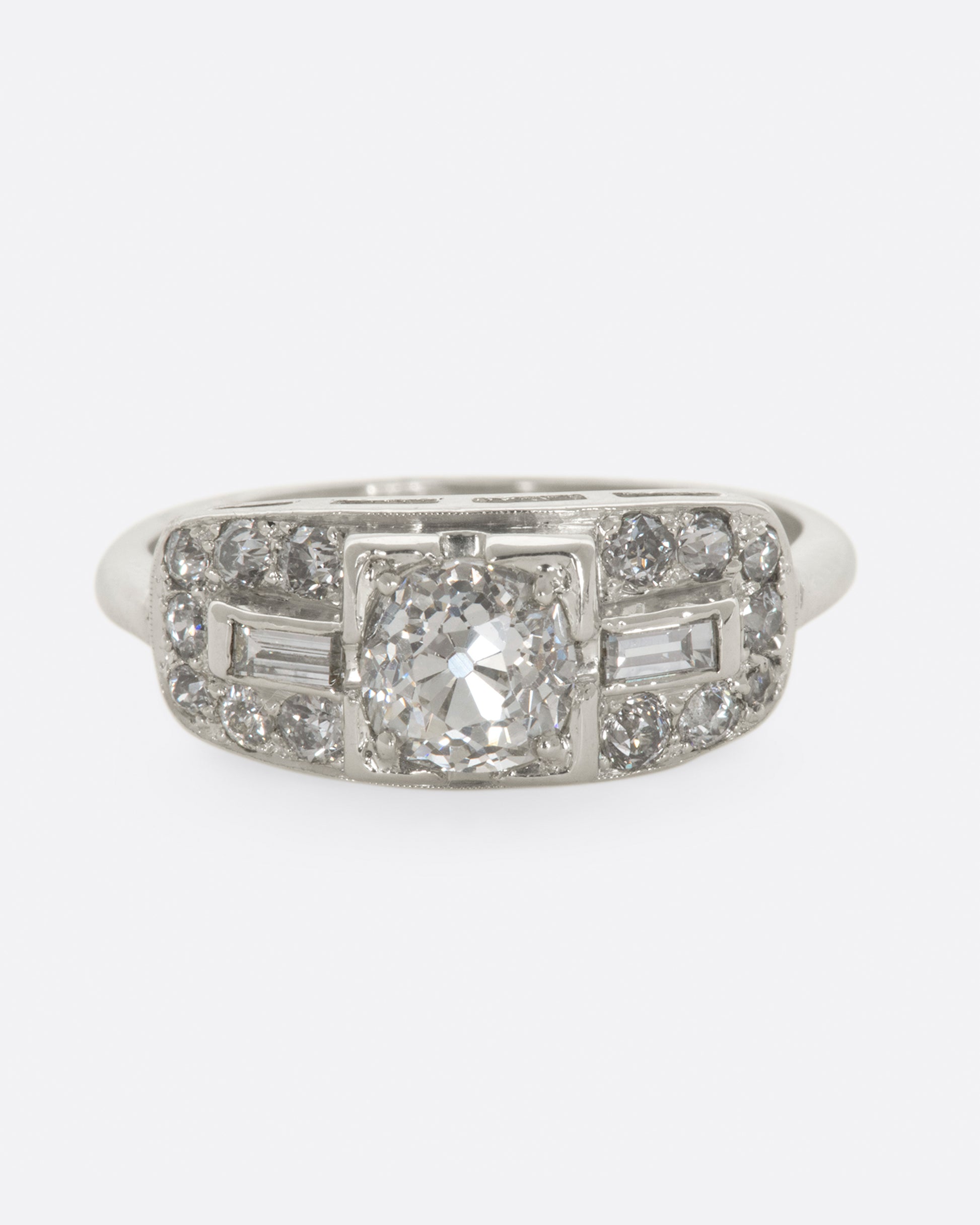 A white gold ring with round center diamond and baguette and round diamonds surrounding on a wide flat face. View from the front.