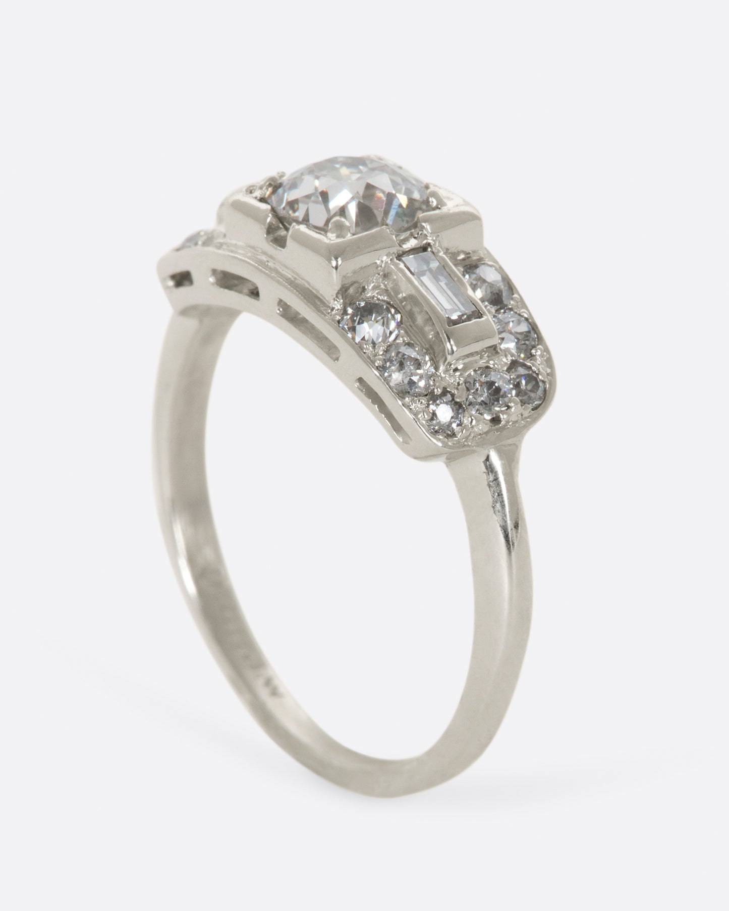 A white gold ring with round center diamond and baguette and round diamonds surrounding on a wide flat face. View standing up.