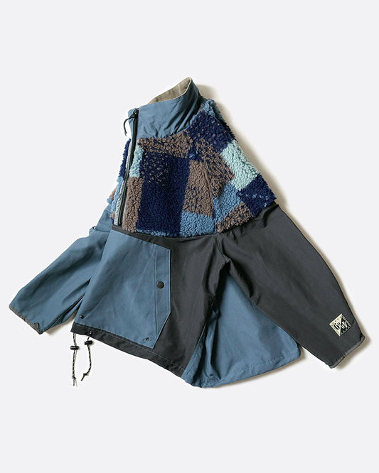 A pullover anorak with a half zip. Half blue and half gray with patchwork fleece around the shoulders. Shown laying flat, but from the side.