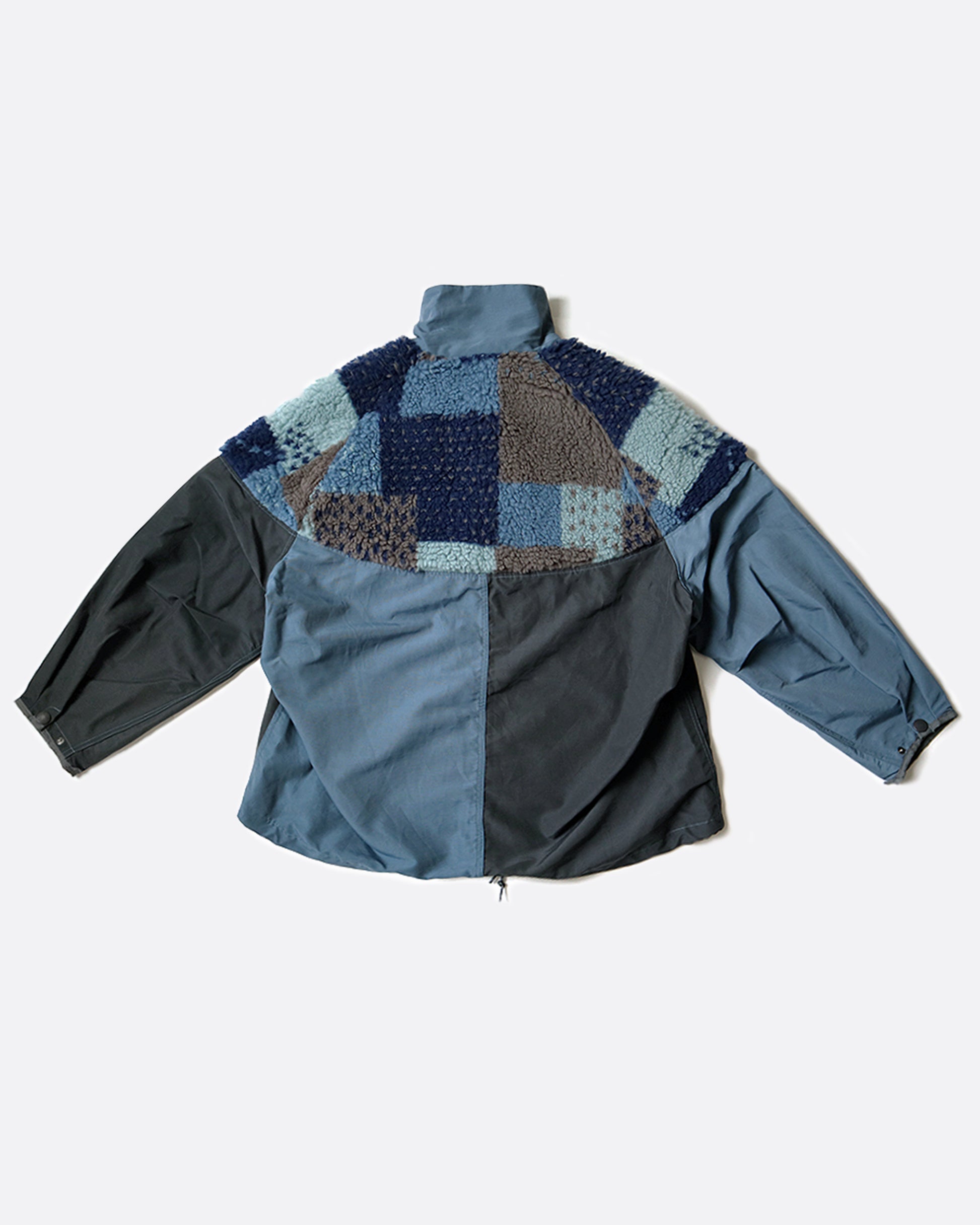 A pullover anorak with a half zip. Half blue and half gray with patchwork fleece around the shoulders. Shown laying flat from the back.