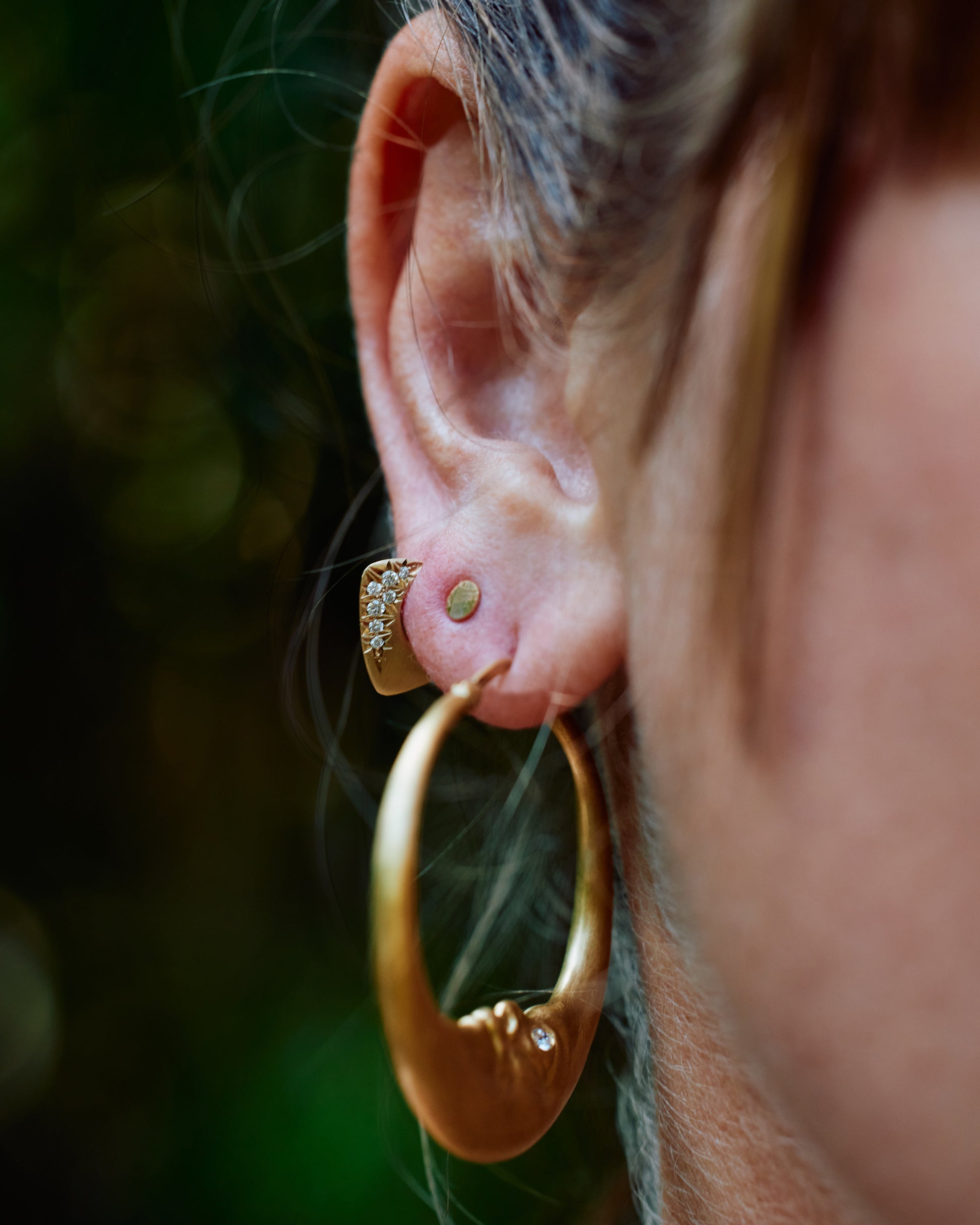 An ear with three earrings; a large gold moon hoop, a round disk stud, and a Yemaya hoop.