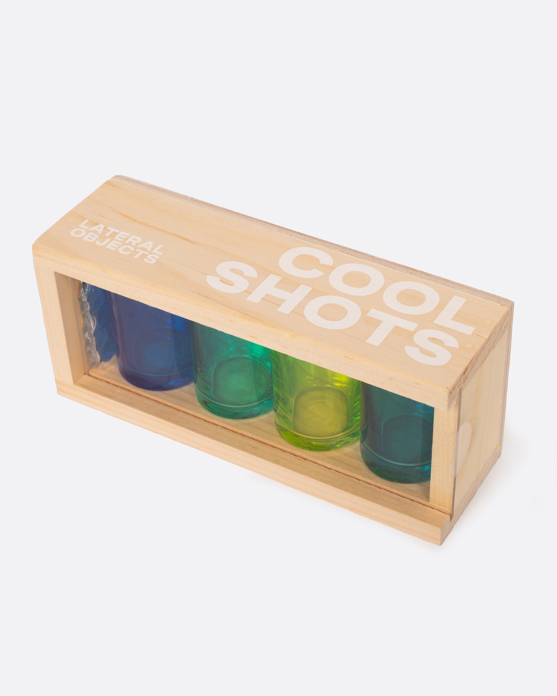 Four glass shot glasses in shades of green and blue, shown in their wood carrying case.
