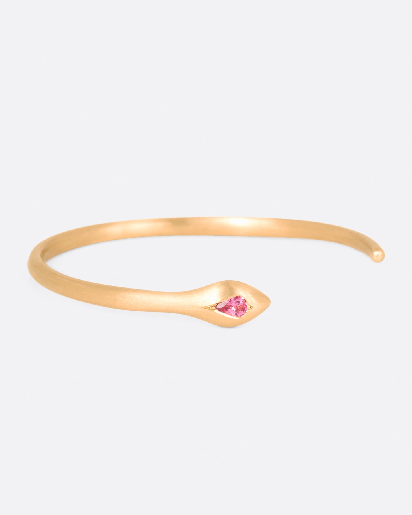 A matte yellow gold snake cuff bracelet with a pear shaped pink sapphire on its head, shown from the front.