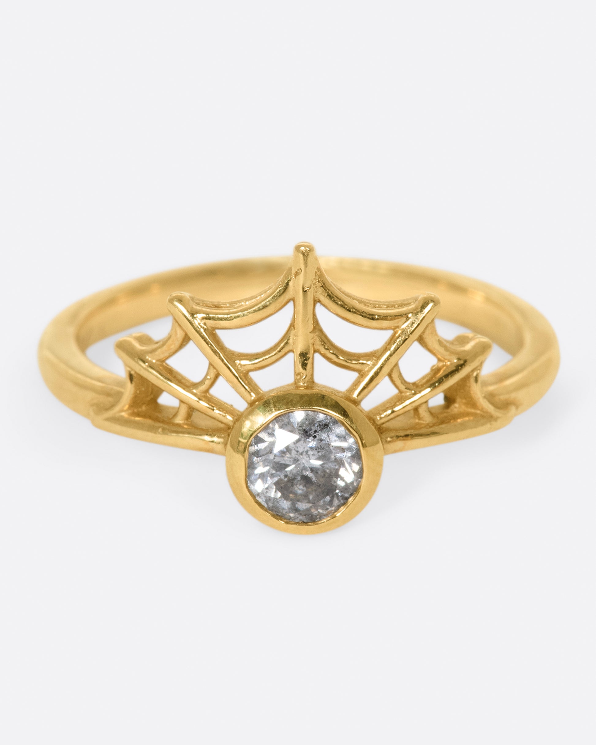 in a yellow gold witchy ring, a grey diamond is bezel set in the middle, with an arched spider web on one side of the diamond.