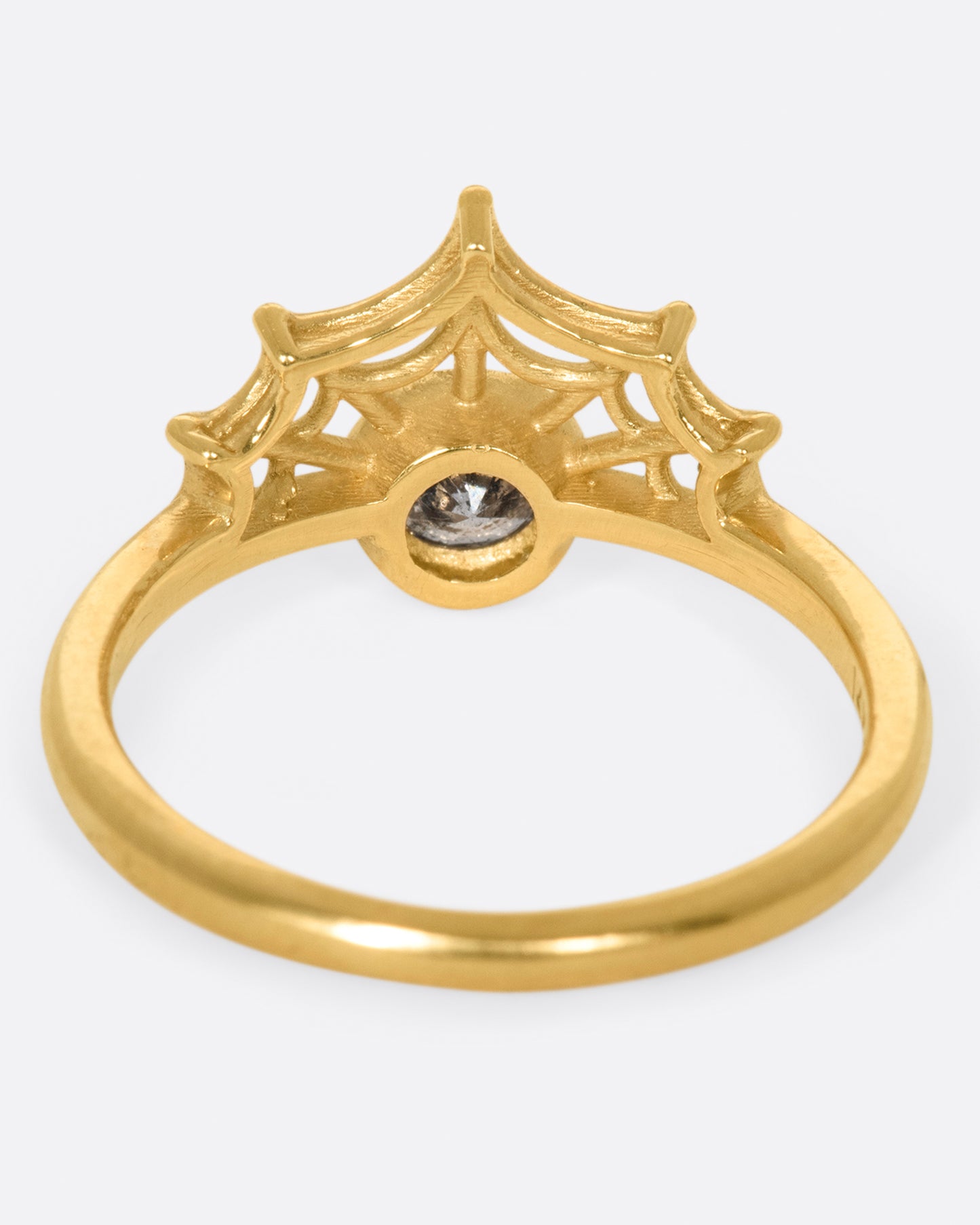 in a yellow gold witchy ring, a grey diamond is bezel set in the middle, with an arched spider web on one side of the diamond.