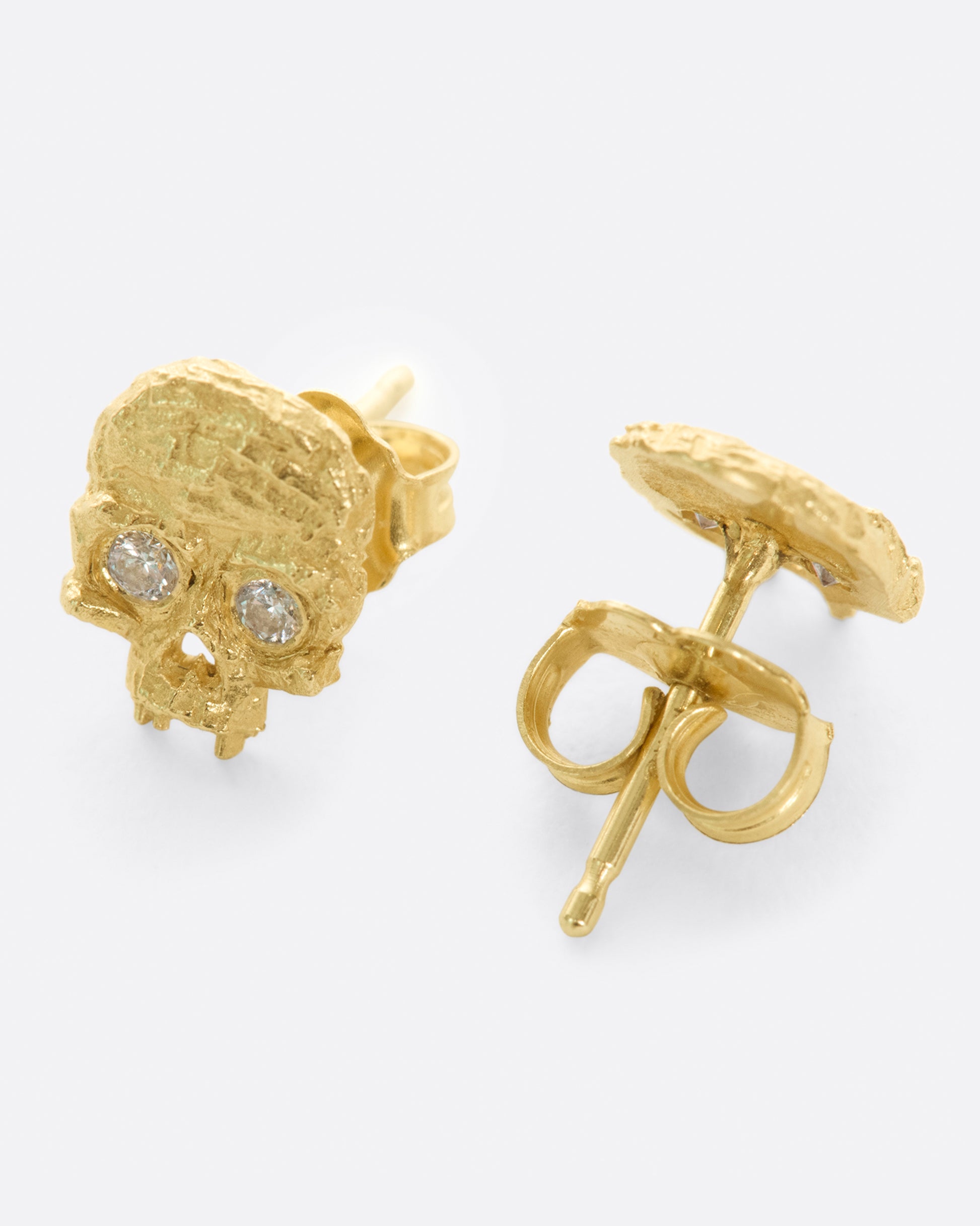 A pair of yellow gold textured skull stud earrings with diamond eyes, shown from the side.