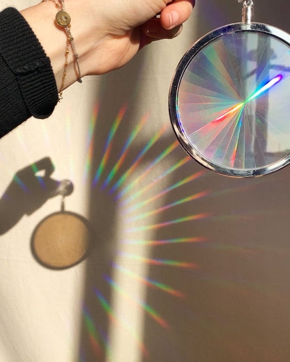 A hand holding a circular rainbow maker with a silver edge and suction cup at the top. The shadow of the hand and maker and many rainbows are shown on the wall in the background. Shown from the front.