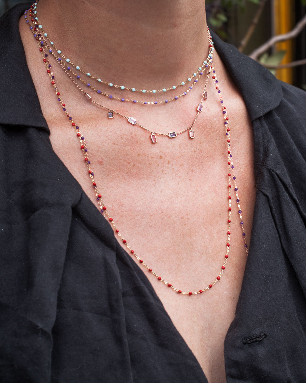 Closeup of woman's neck; she wears multiple yellow gold chain necklaces with resin beads of various lengths in addition to another necklace with precious gemstones and a black shirt. 