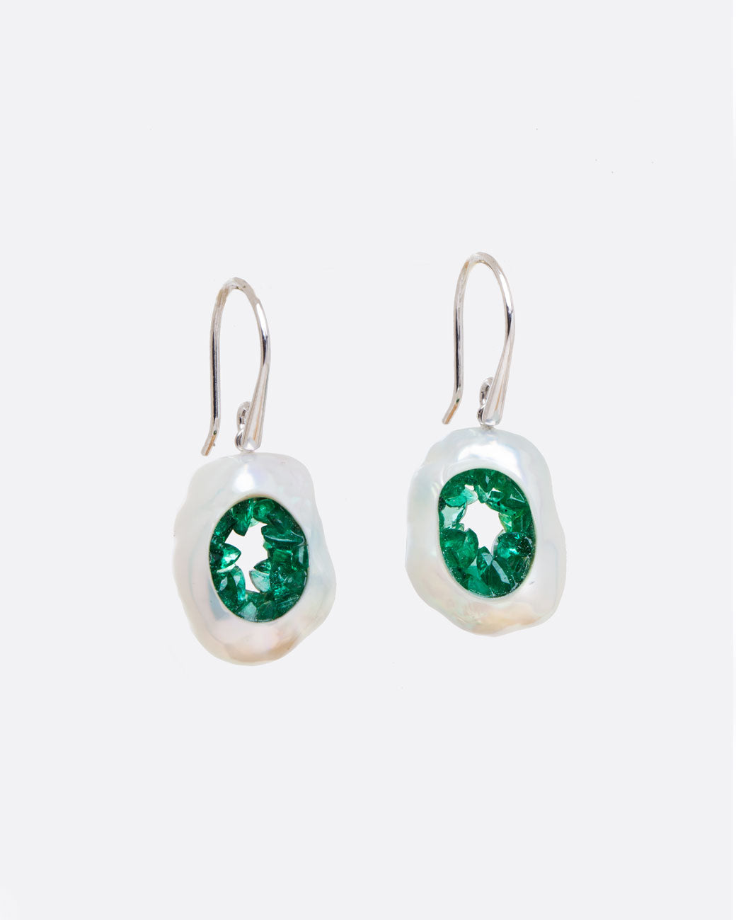 two dangle hook earrings in white gold hanging and viewed from the front. the dangle part is a pearl that has had a hole carved out in the middle and then lined with emeralds, to create a geode like appearance.