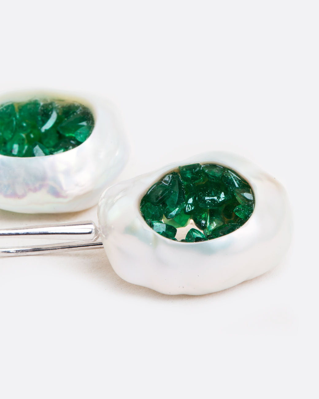 two dangle hook earrings in white gold laying on a white surface. the dangle part is a pearl that has had a hole carved out in the middle and then lined with emeralds, to create a geode like appearance.