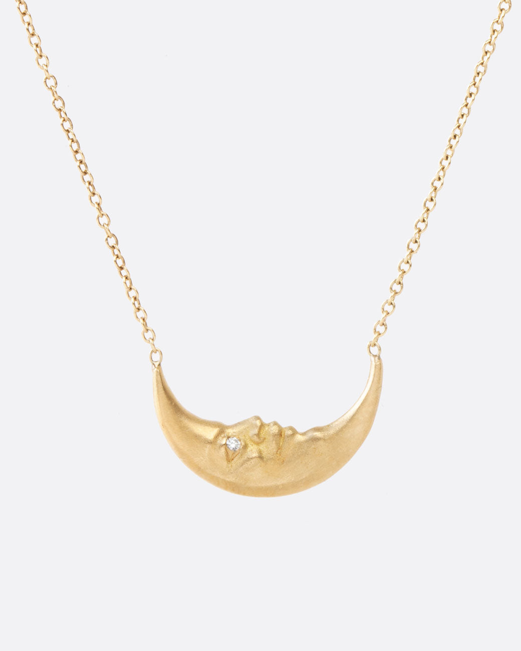 Super close up view of a tiny yellow gold crescent moon, with a face carved into it and little diamond eyes, is a unique pendant. Double sided and hanging from a chain. The chain is attached to the ends of the crescent, so it falls on a sideways/horizontal angle across the chest.