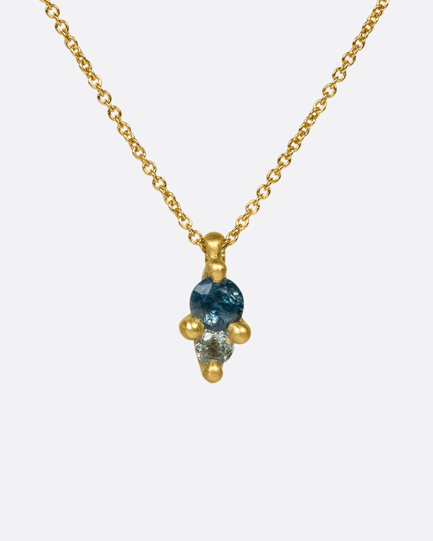 A pendant with two sapphires; one blue and one gray, set in matte gold prongs on a cable chain necklace.