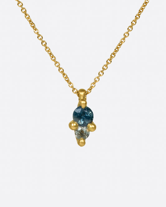 A pendant with two sapphires; one blue and one gray, set in matte gold prongs on a cable chain necklace.