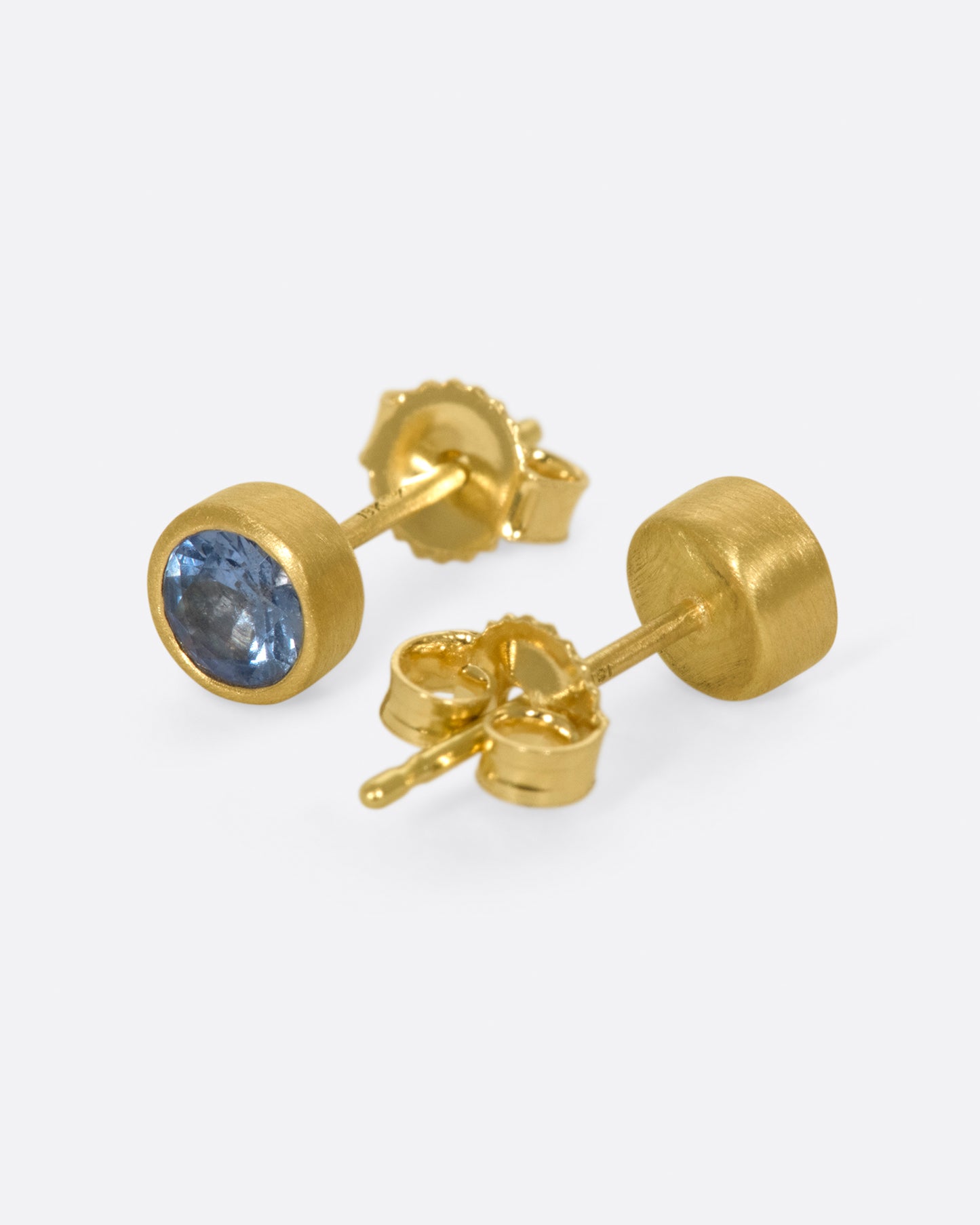A pair of solitaire earrings featuring medium-blue sapphires in matte gold bezels.
