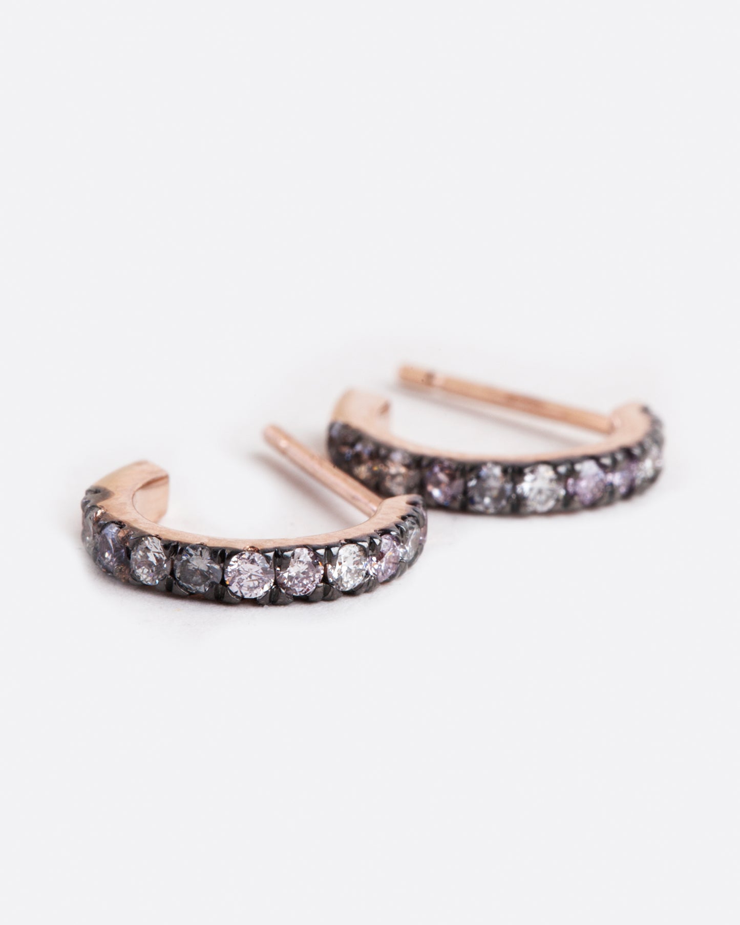 side view of huggie hoop earrings with a stud post backing, they are rose gold with blackened settings around the grey, pink and white diamonds that are set along the huggie