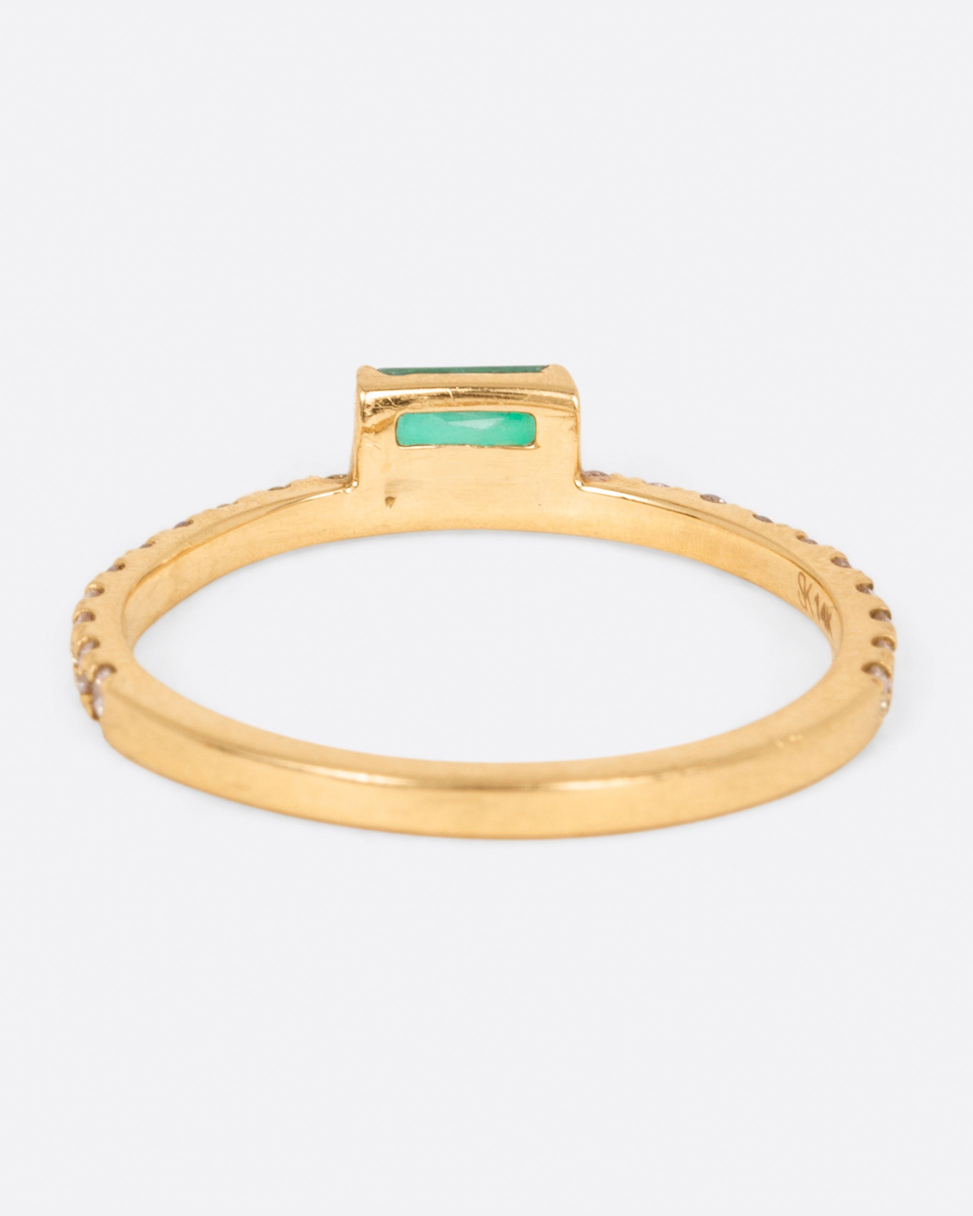 A yellow gold ring with white diamonds going 2/3 of the way around and an emerald baguette sitting atop it, shown from the back.