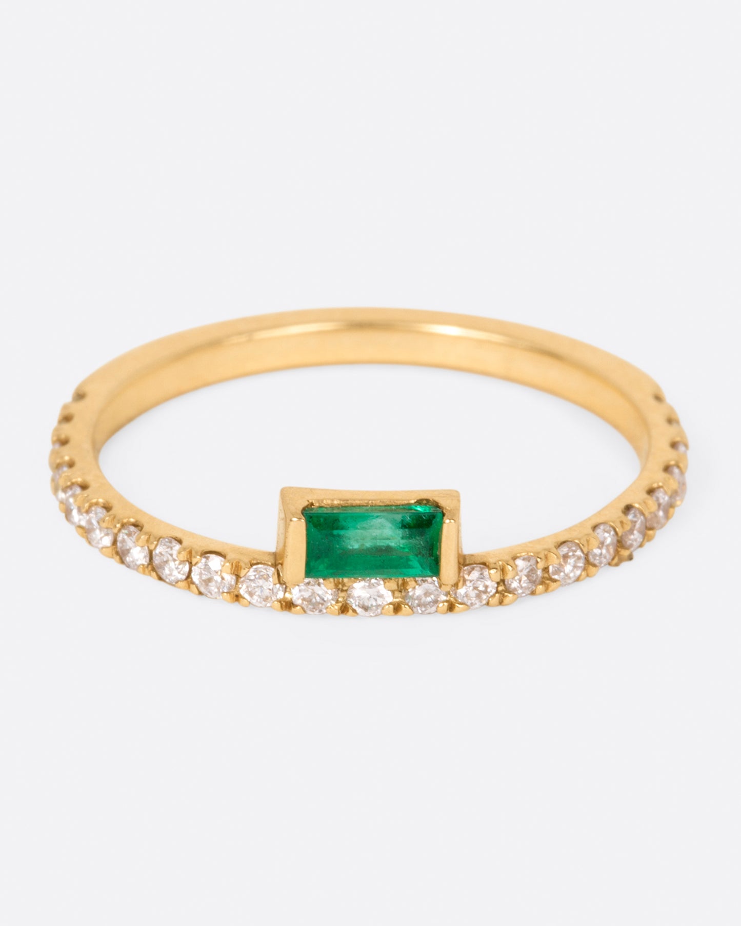 A yellow gold ring with white diamonds going 2/3 of the way around and an emerald baguette sitting atop it, shown from the front.