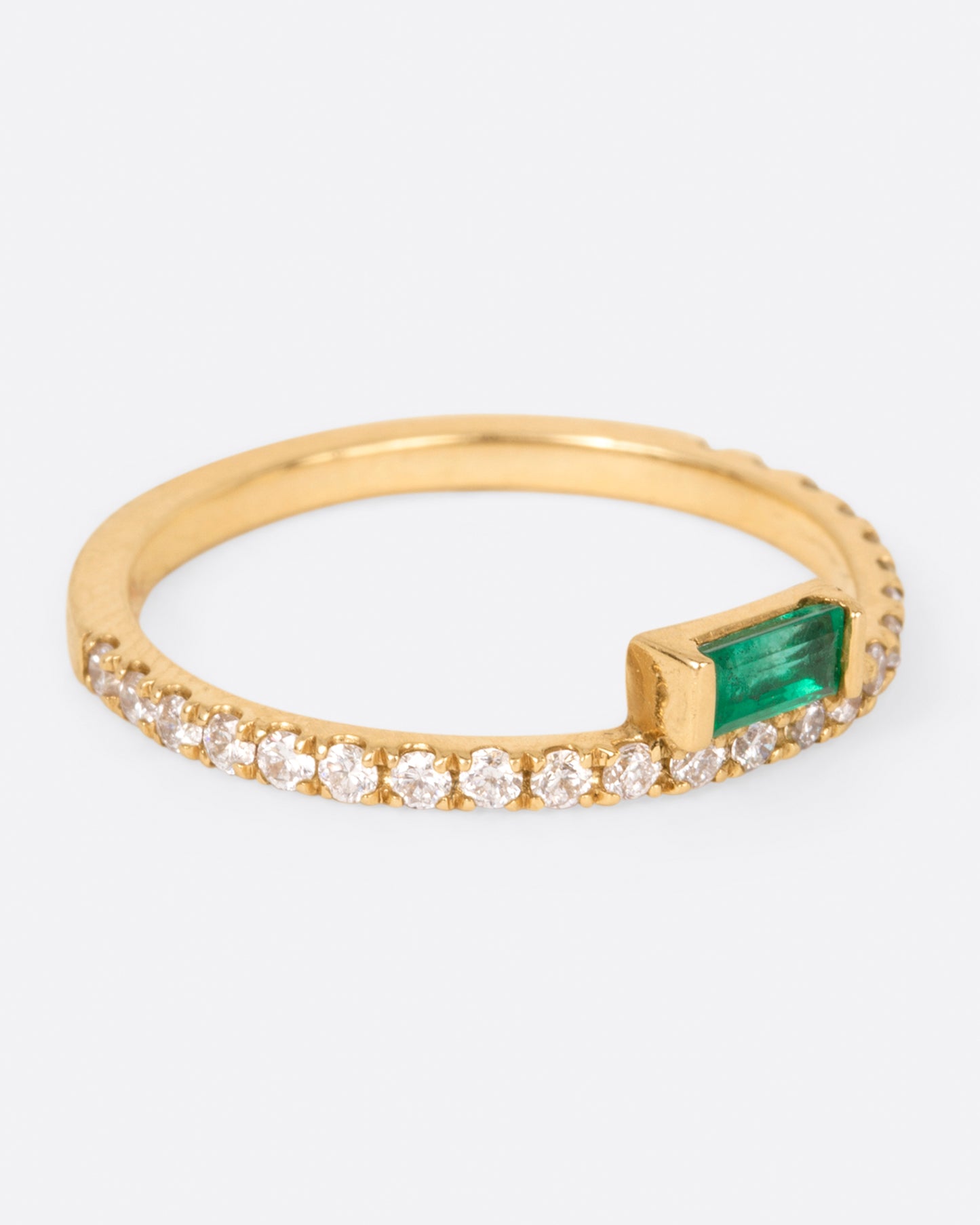 A yellow gold ring with white diamonds going 2/3 of the way around and an emerald baguette sitting atop it, shown from the side.